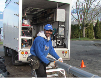 A smiling worker in a blue hoodie and cap handles a large metal pipe near the back of a work truck on a street, with tools and equipment visible and orange safety cones nearby.